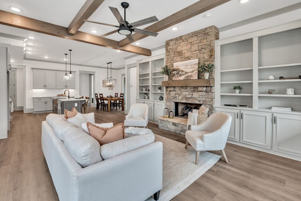 Open-concept living room featuring wood ceiling beams, stone fireplace, and view into modern kitchen and dining area.